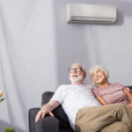 An older couple smile from their couch beneath their air vent.