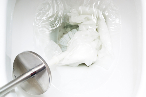 Close-up image of a toilet bowl with multiple wet wipes clogged and a toilet brush trying to push them down