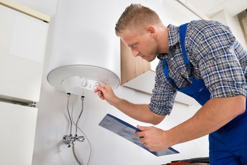 A man wearing a plaid shirt and blue apron holds a clipboard and leans forward to inspect a white tankless water heater.