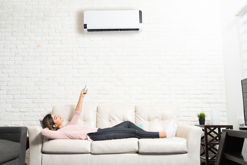 A woman lays on the couch and points her remote control at her mini split to adjust the temperature. 