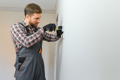 An electrician wearing a plaid shirt and gray overalls screws a panel into a white wall.